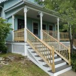 “Blue suburban house with a white-trimmed porch, wooden staircase, and intricate balusters, surrounded by lush greenery, showcasing XTEND Contracting’s expertise in porch and deck construction.